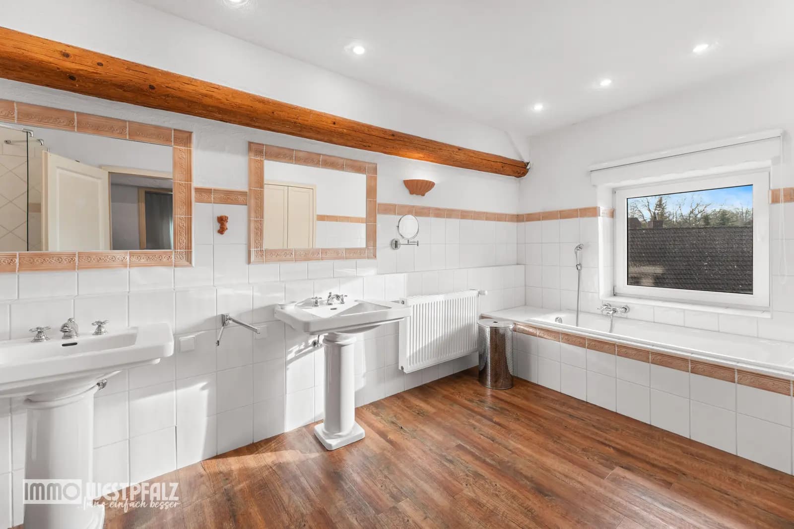 Helles Badezimmer mit Doppelwaschbecken, Badewanne am Fenster und rustikalem Holzbalken — professionelle Immobilienfotografie.