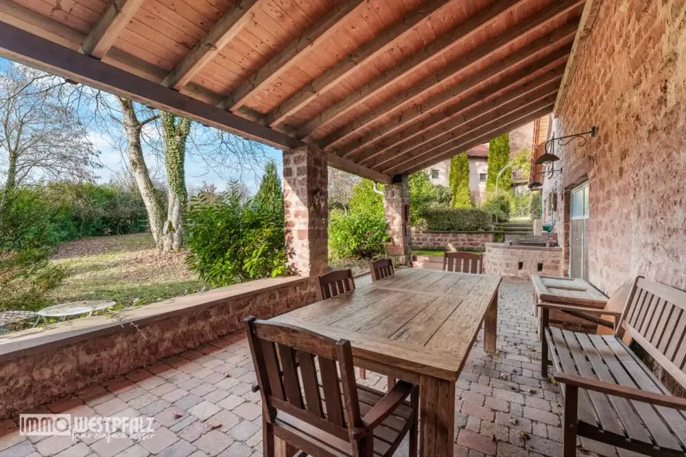 Überdachte Terrasse mit Sandsteinmauerwerk, großem Holztisch und Blick in den Garten — Außenaufnahme für die Immobilienvermarktung.