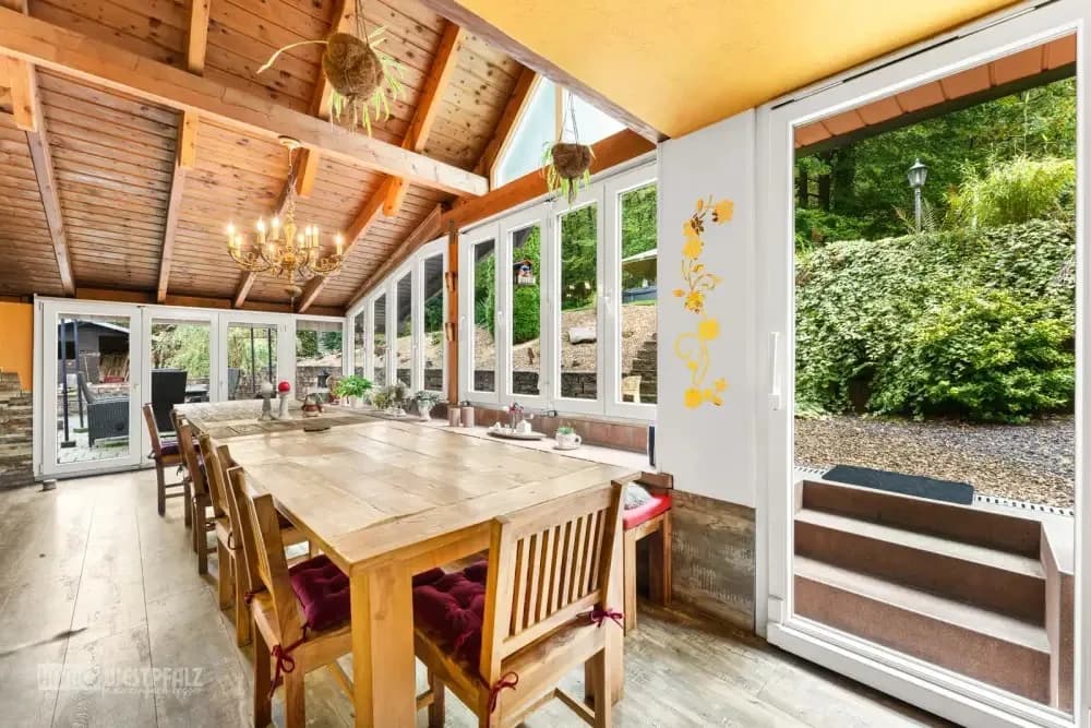 Heller Wintergarten mit großem Holzesstisch, rustikaler Holzdecke und Blick in den grünen Garten — Immobilienaufnahme in der Südwestpfalz.