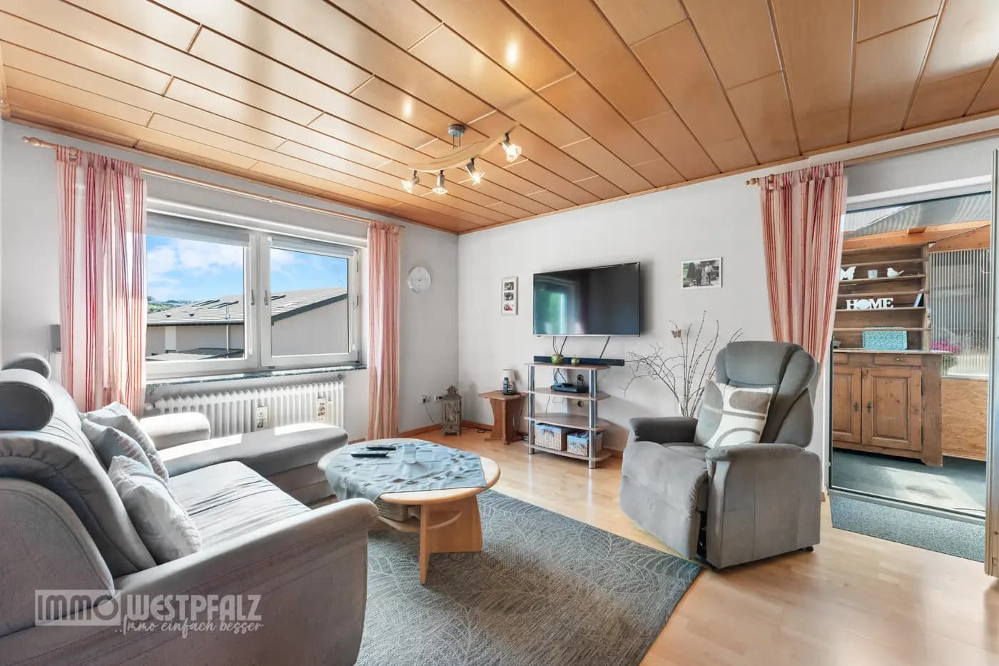 Freundliches Wohnzimmer mit Holzdeckenpaneelen, grauer Eckcouch und großem Fenster — natürliches Licht in der Immobilienaufnahme.